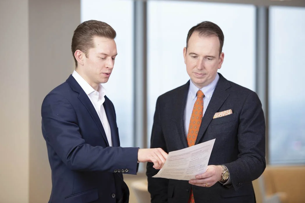Two professionally dressed men in suits look at a document together in an office setting.