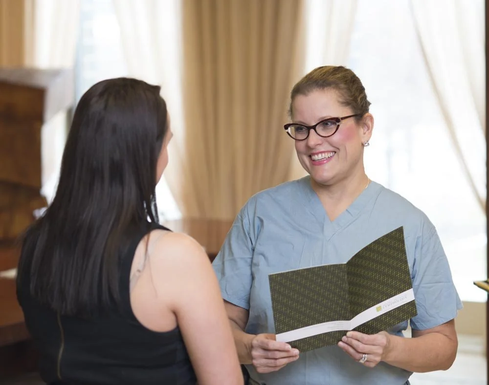 A woman in medical scrubs smiling and holding a brochure, talking to a woman with long dark hair.