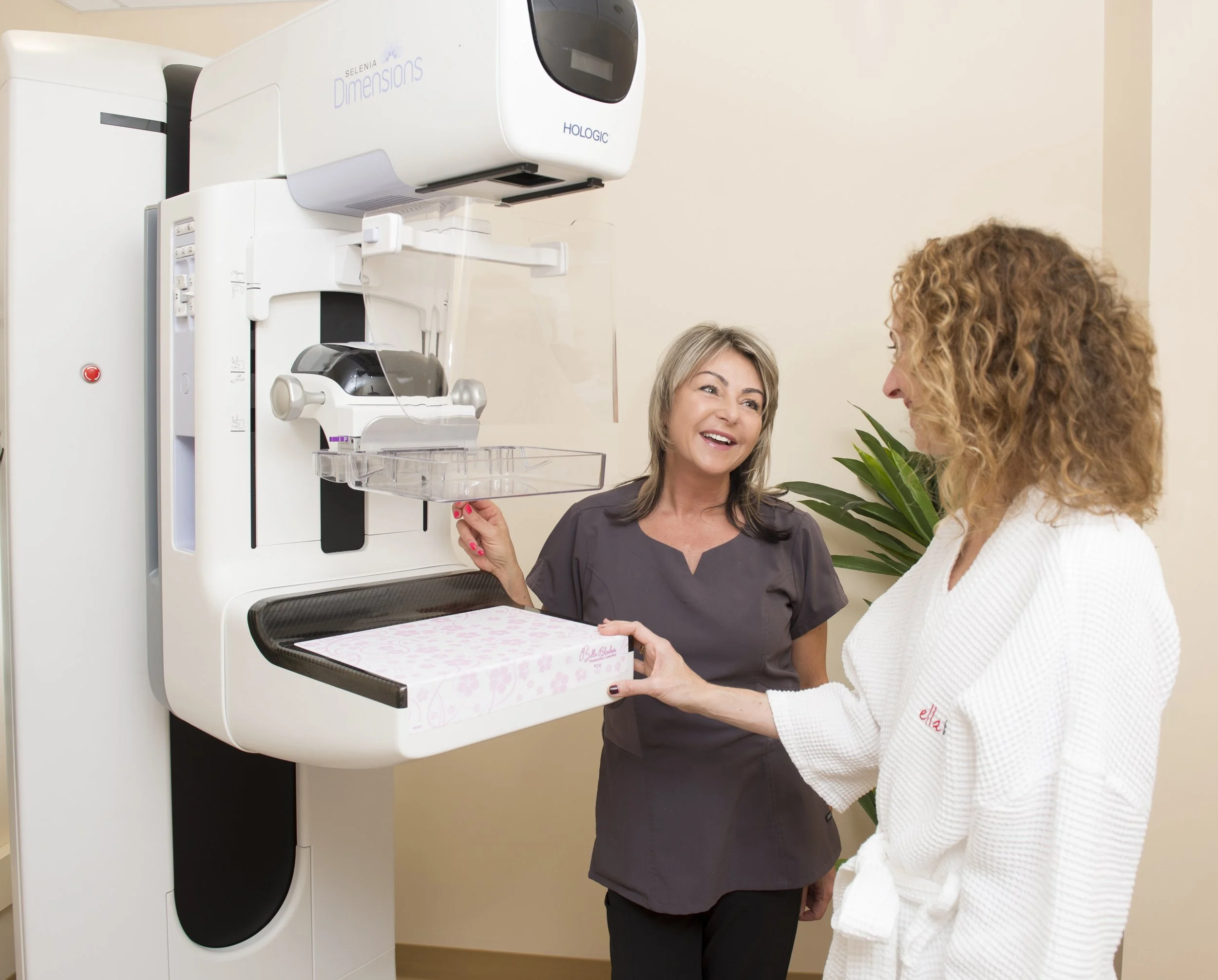 A woman in a white robe is handing a pink box to a woman in gray scrubs in front of a mammogram machine in a medical clinic.