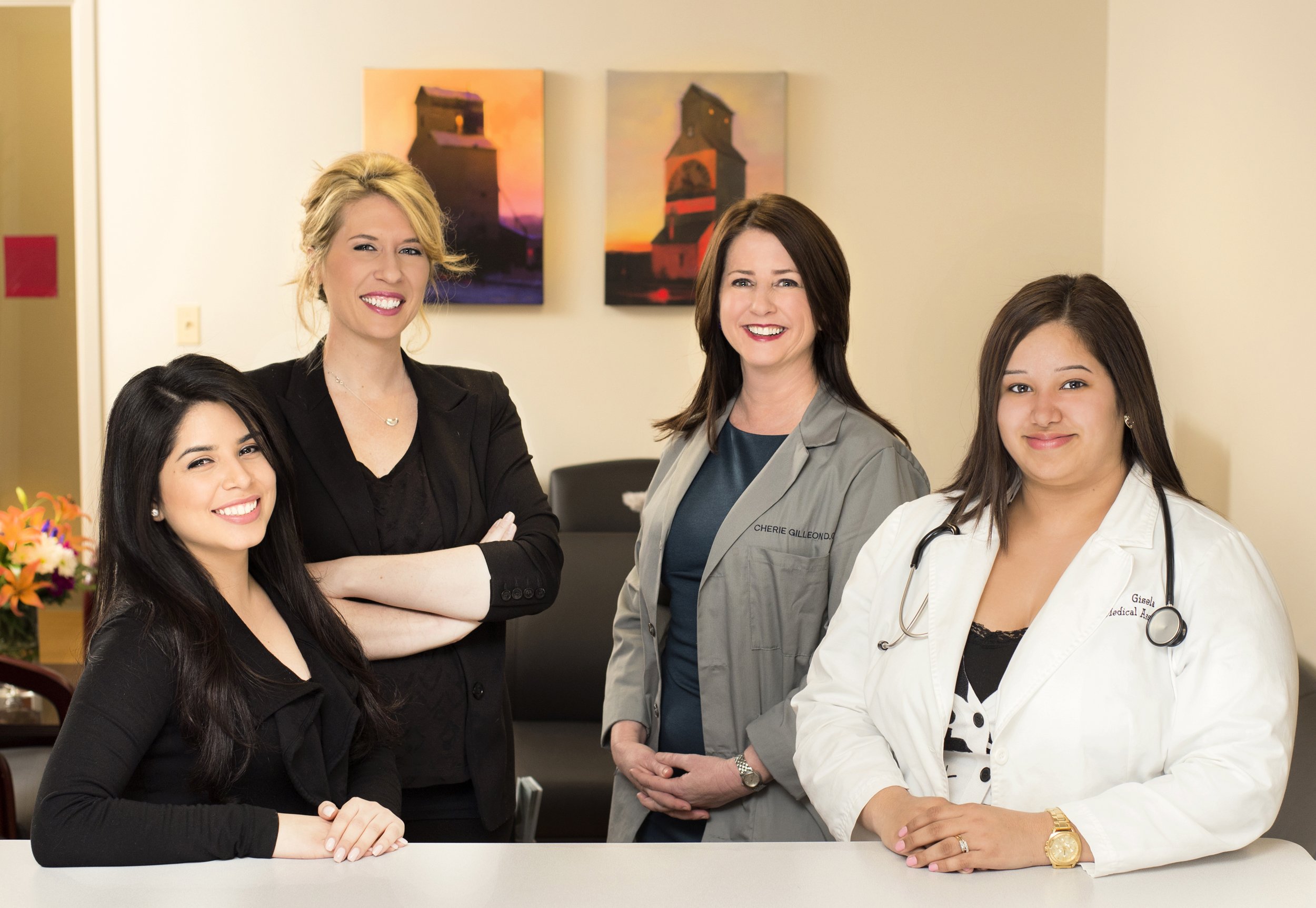 Four women, including two healthcare professionals in white coats with stethoscopes, smiling in a medical office or clinic setting.