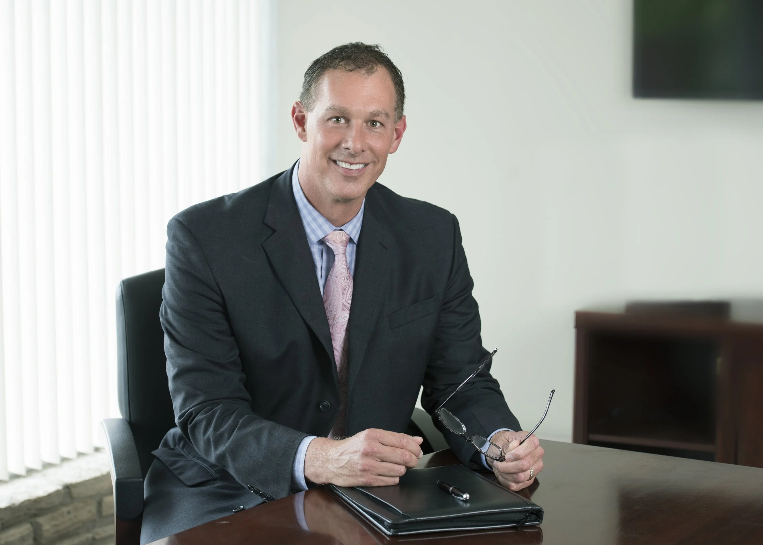 Chicago portrait photography of a wealth manager. The man is sitting at his desk with a notebook and glasses with his office as the environment blurred out of focus. This photo is of the CEO and owner of the firm and will be used for annual reports. 