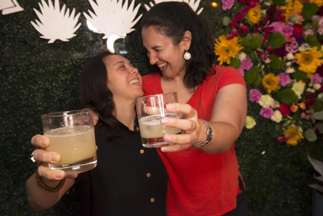 Two women celebrating, smiling and holding drinks, with a floral and decorative background.