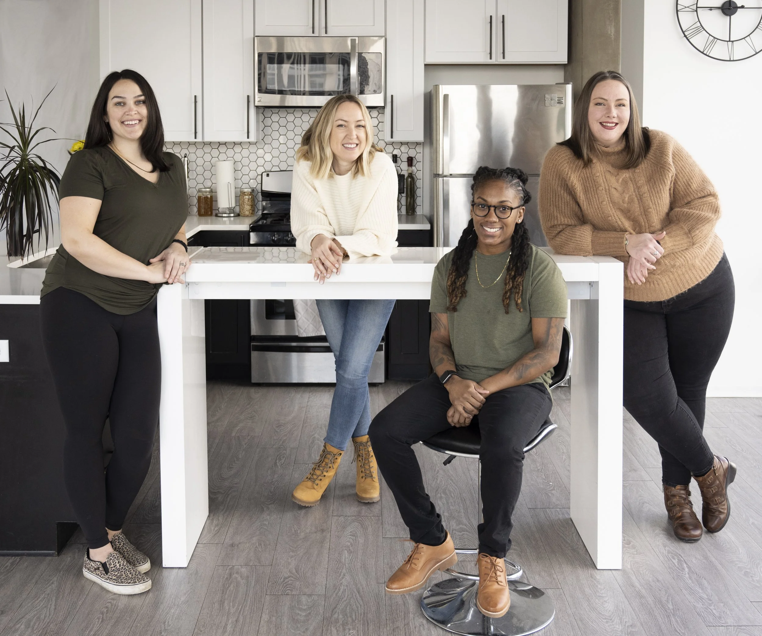 Four women smiling in a modern kitchen, one sitting on a stool and three standing behind a white kitchen island.
