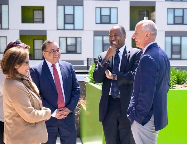 Four business professionals, two men and two women, are standing outside, engaged in conversation and laughing, with a modern apartment building in the background.