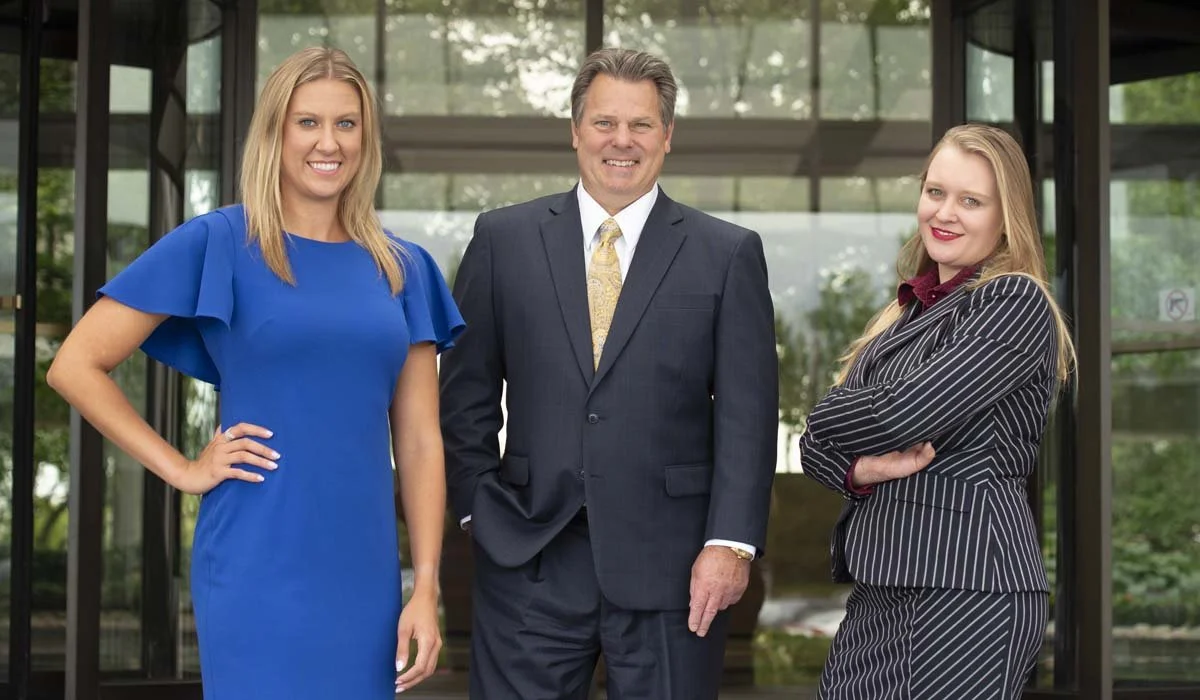 Three professionally dressed individuals, two women and one man, standing outside a glass building with greenery in the background. The woman on the left wears a blue dress, has blonde hair, and is smiling with her hand on her hip. The man in the mid