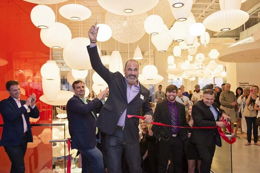 Man with beard in suit cutting a red ribbon at an event, surrounded by people, with hanging paper lanterns overhead.