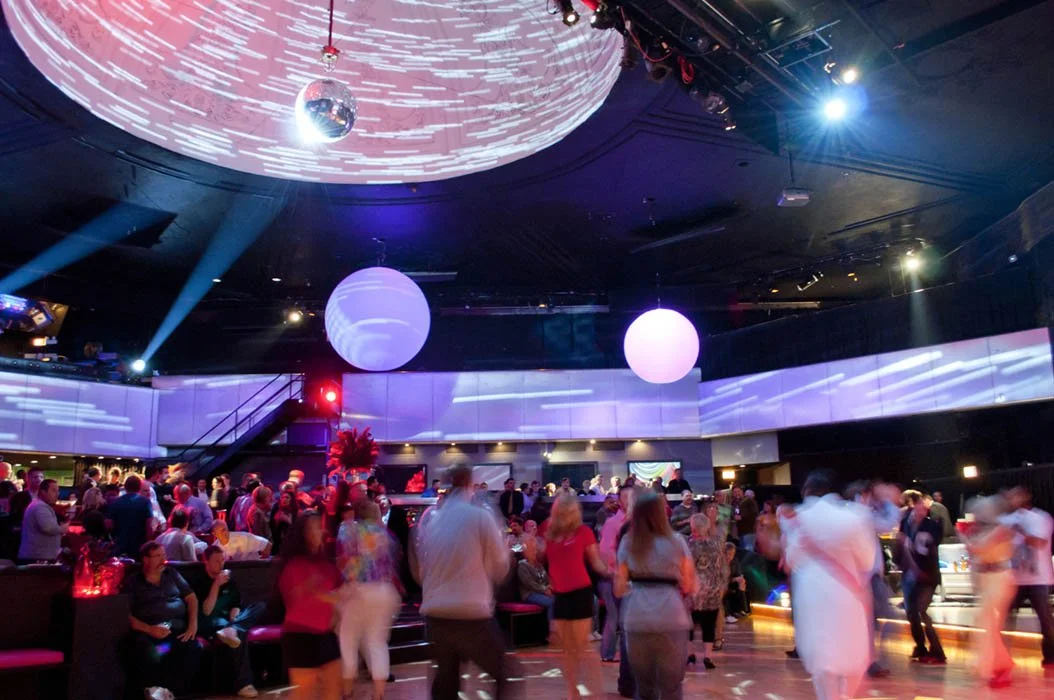 People dancing and socializing inside a nightclub with colorful lighting, large white spherical lamps, a disco ball on the ceiling, and a DJ booth in the background at Park West