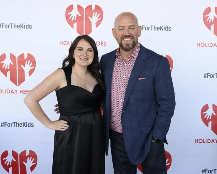 A woman in a black dress and a man in a blue suit with a pink shirt are smiling and posing together in front of a step and repeat backdrop with a red broken heart logo.
