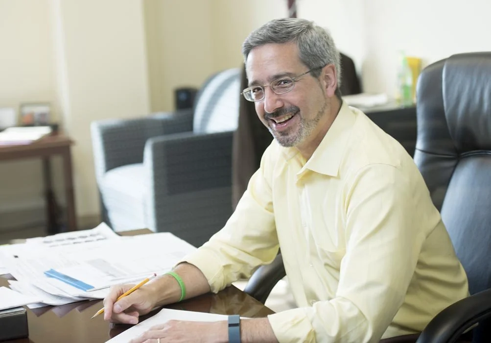 A middle-aged man with glasses, a beard, and gray hair, smiling, sitting at a desk cluttered with papers, holding a pencil, in an office setting.