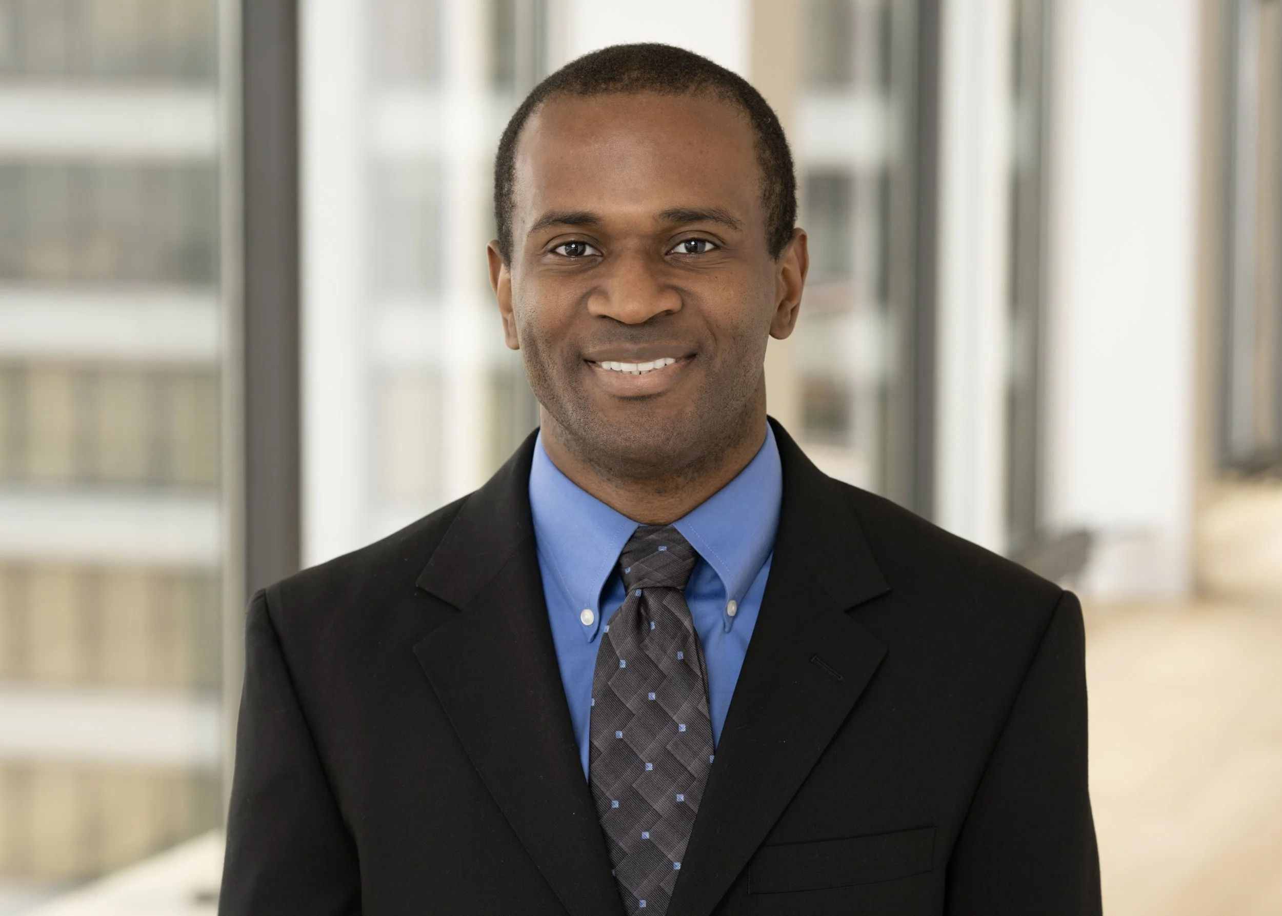 Professional headshot of an attorney in a suit standing in a modern office building with large windows with the Chicago skyline out of focus.