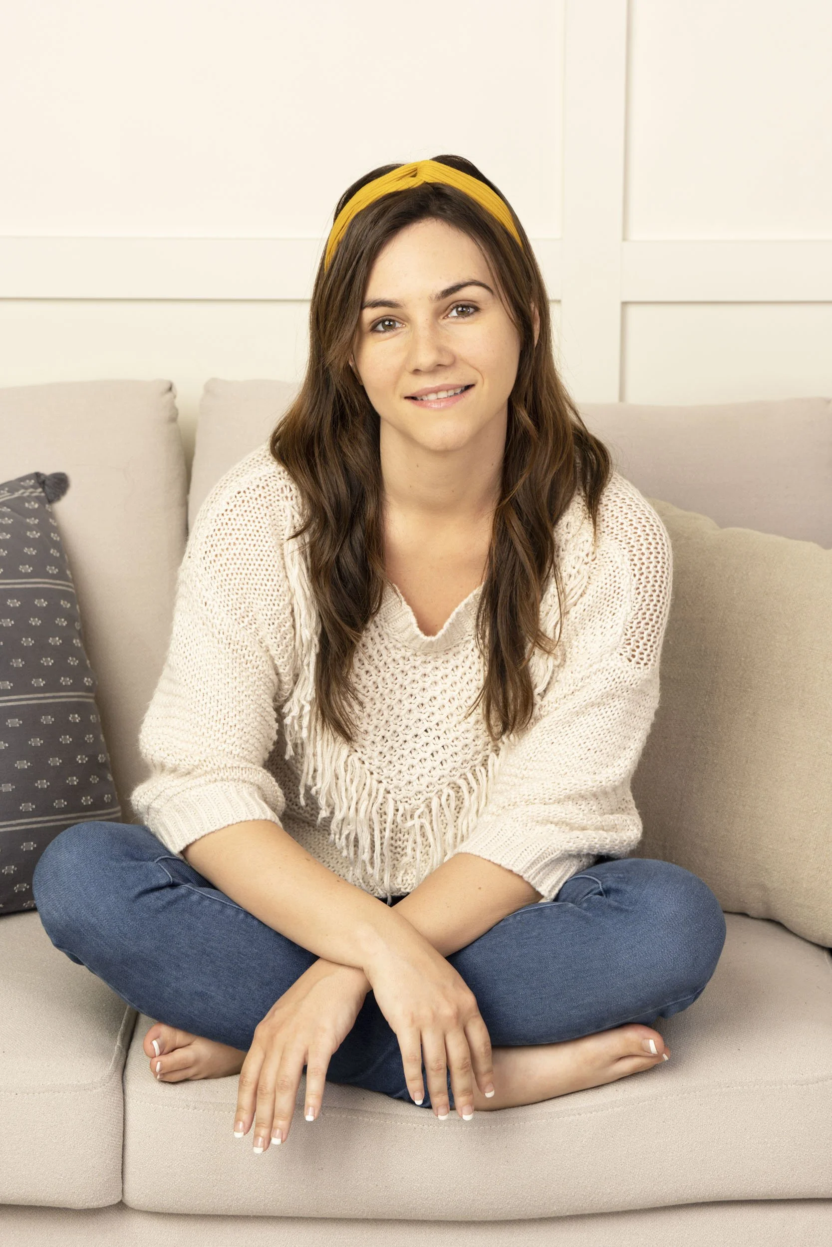 Portrait photography of an young woman influencer with long brown hair, yellow headband, wearing a cream knit sweater and blue jeans, sitting cross-legged on a beige sofa in a cozy room.