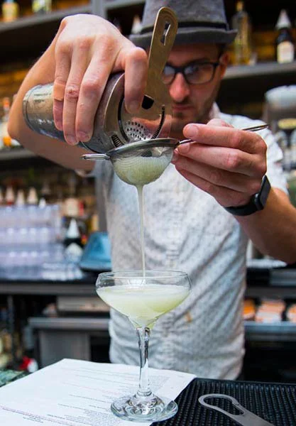 A bartender straining a light green cocktail into a coupe glass through a fine-mesh sieve in a bar or restaurant.