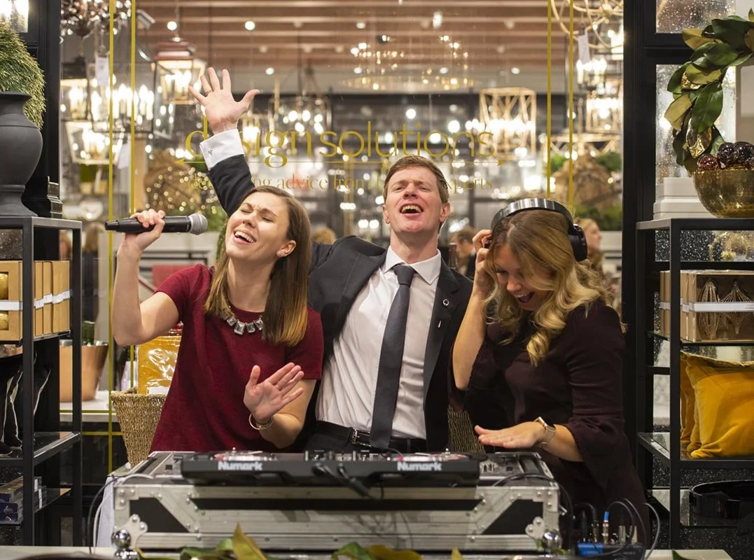 Three people joyfully singing and dancing around a DJ booth at a party or celebration inside a store during the grand opening, with decorative shelves and plants in the background.
