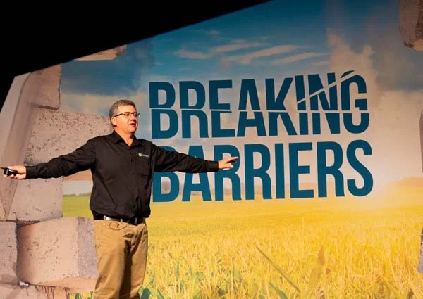 A man in a black shirt and khaki pants giving a presentation with his arms outstretched in front of a sign that reads 'Breaking Barriers' in bold blue letters and a background of a field and blue sky.