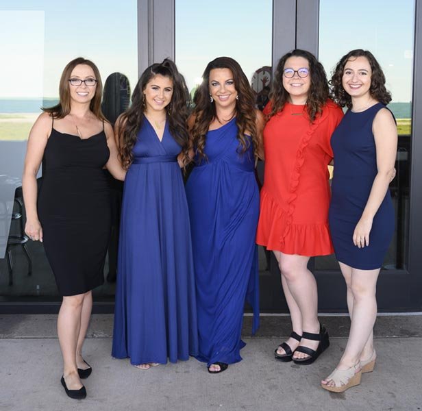 Group photo of five women standing together outdoors in front of glass doors, smiling, dressed in colorful dresses, with a scenic view of water and green fields in the background at a company party.