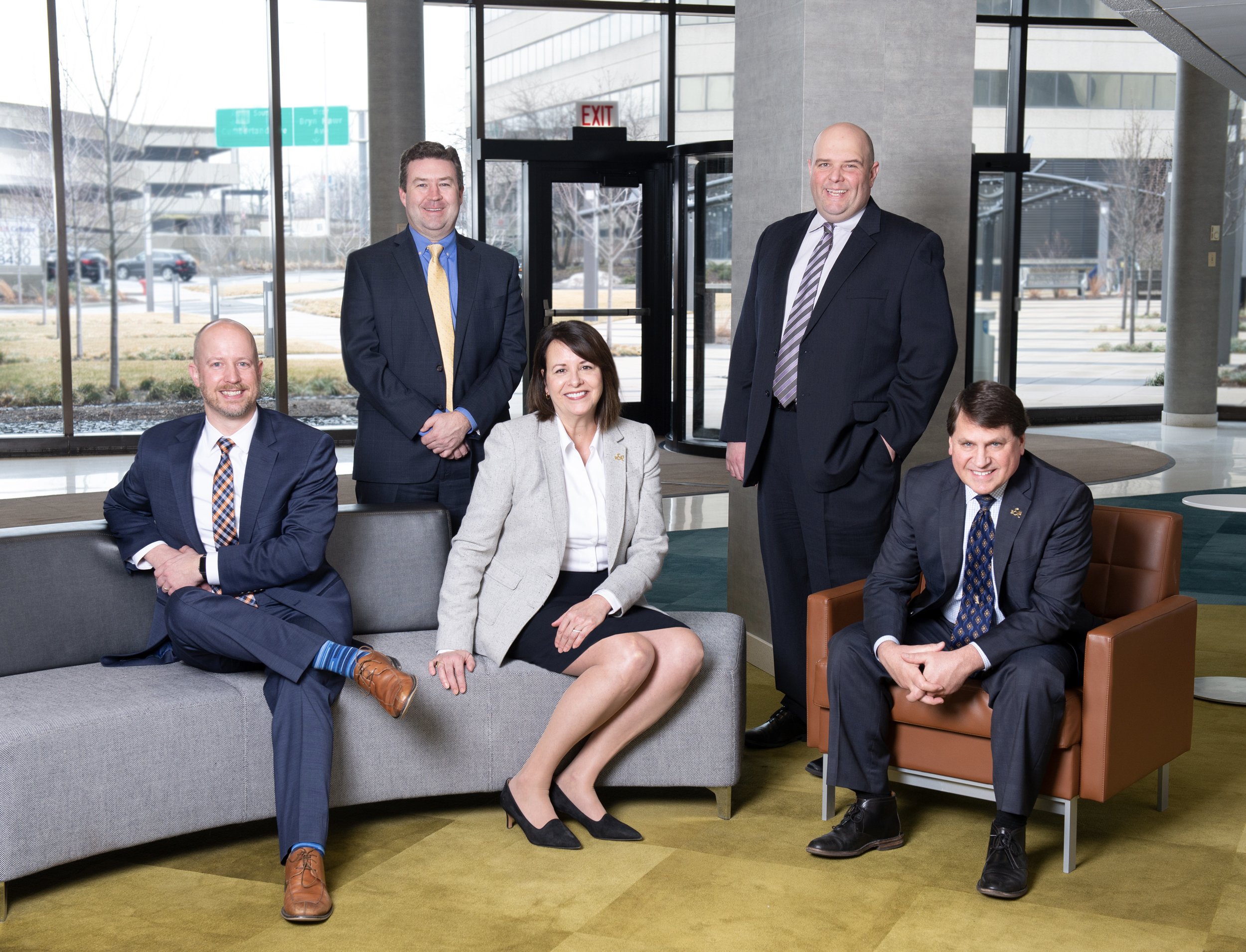 Five business professionals, four men and one woman, posing for a group photo in a modern office lobby with large glass windows and glass doors in the background. Two men are standing, and three are sitting on sofas and armchairs, dressed in formal b