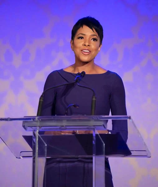 A woman with short black hair wearing a dark-colored dress is speaking at a podium with a microphone, in front of a blue and purple background.