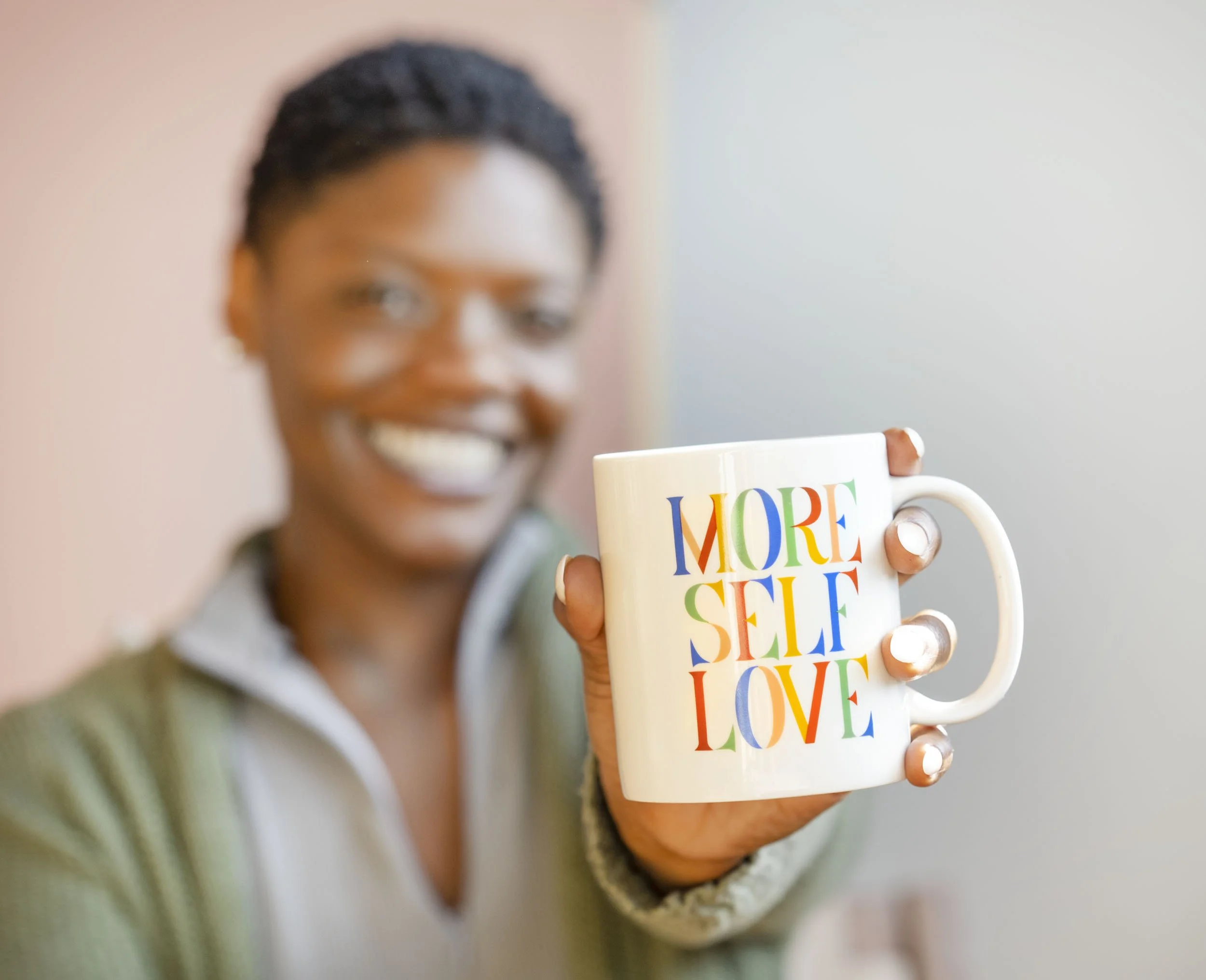 A social media influencer smiling holding a white mug with the words 'MORE SELF LOVE' written in colorful letters.