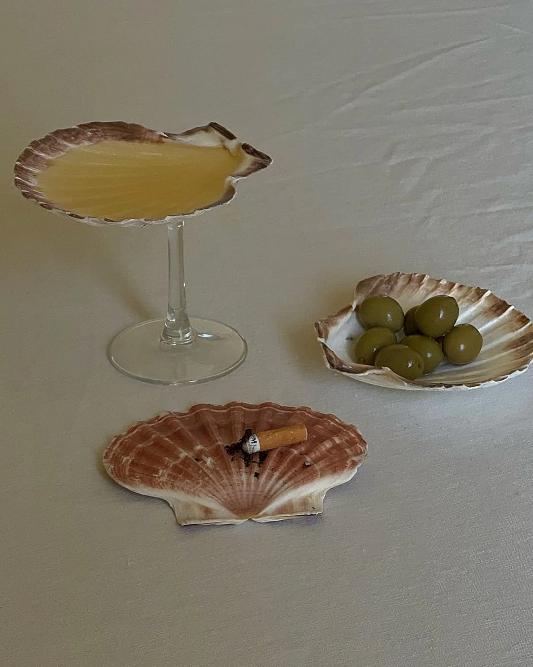 A still life of seashell dishes with a drink glass, green olives, and a cigarette on a light-colored surface.