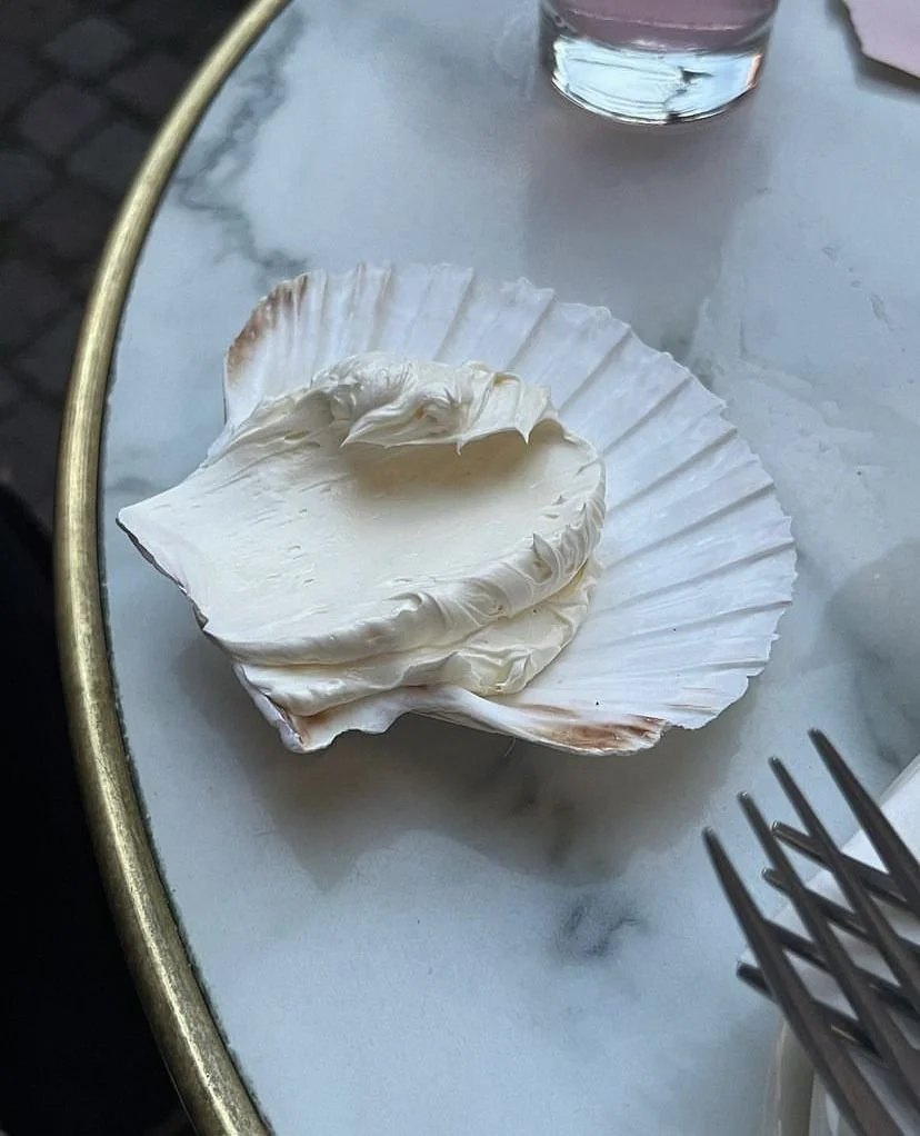 A shell with a serving of creamy, white cheese on a small paper dish, placed on a round table with a glass of water nearby.