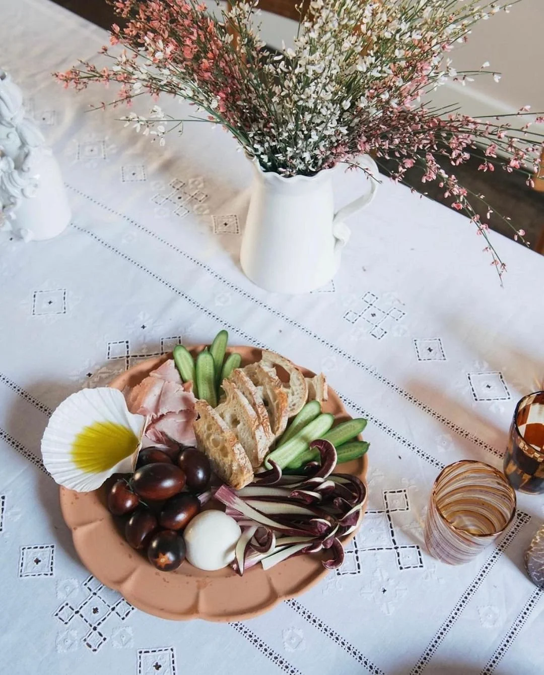 A table with a vase of pink and white flowers, a plate of assorted snacks including grapes, sliced bread, and vegetables, and some glasses.