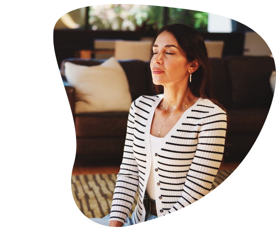 A woman with closed eyes and a peaceful expression, sitting indoors with her hands resting on her lap, wearing a striped blazer and jewelry.