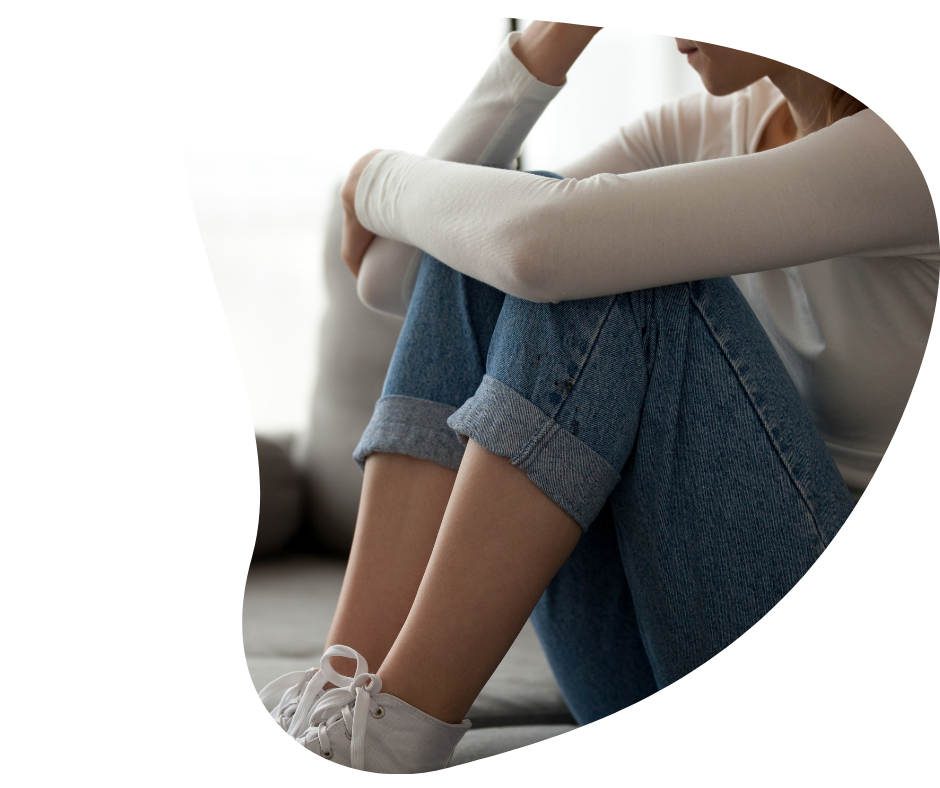 A person sitting on a couch with their knees drawn up, wearing a white long-sleeve shirt, rolled-up blue jeans, and white sneakers.