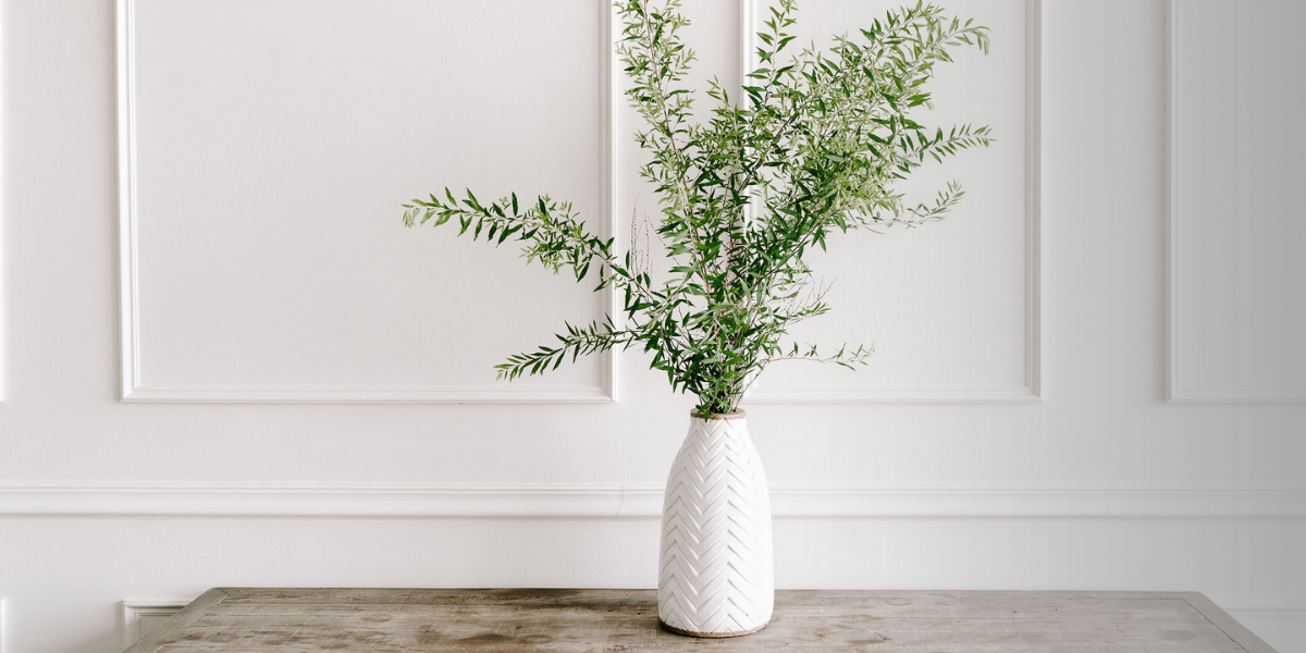 A white ceramic vase with a textured zigzag pattern containing green leafy branches, placed on a wooden surface against a white wall with decorative paneling.