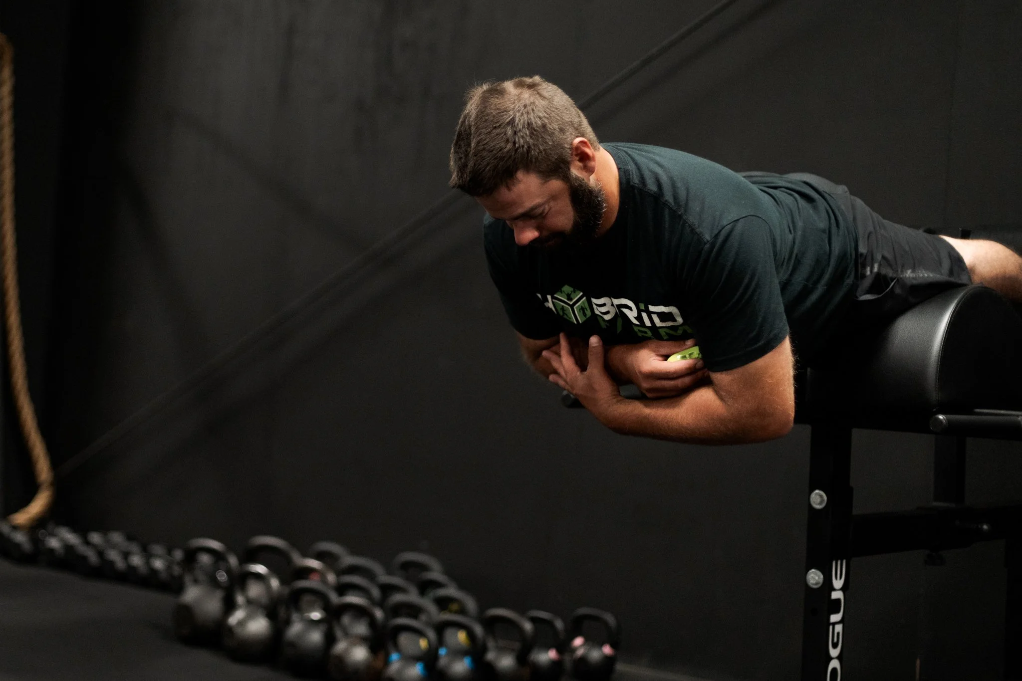 Man with beard performing a plank exercise on a padded bench with kettlebells on the floor next to him, in a gym with black walls.