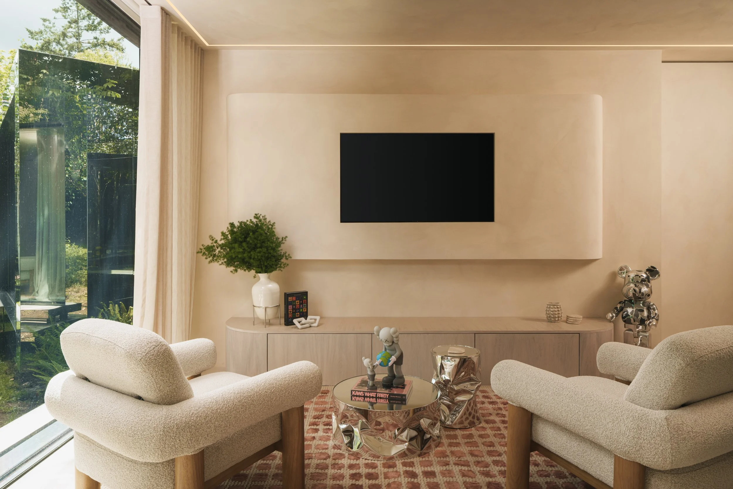 Modern living room painted in limewash with two beige armchairs, a glass-top coffee table, a flat-screen TV on a white wall, and large windows with curtains showing greenery outside.
