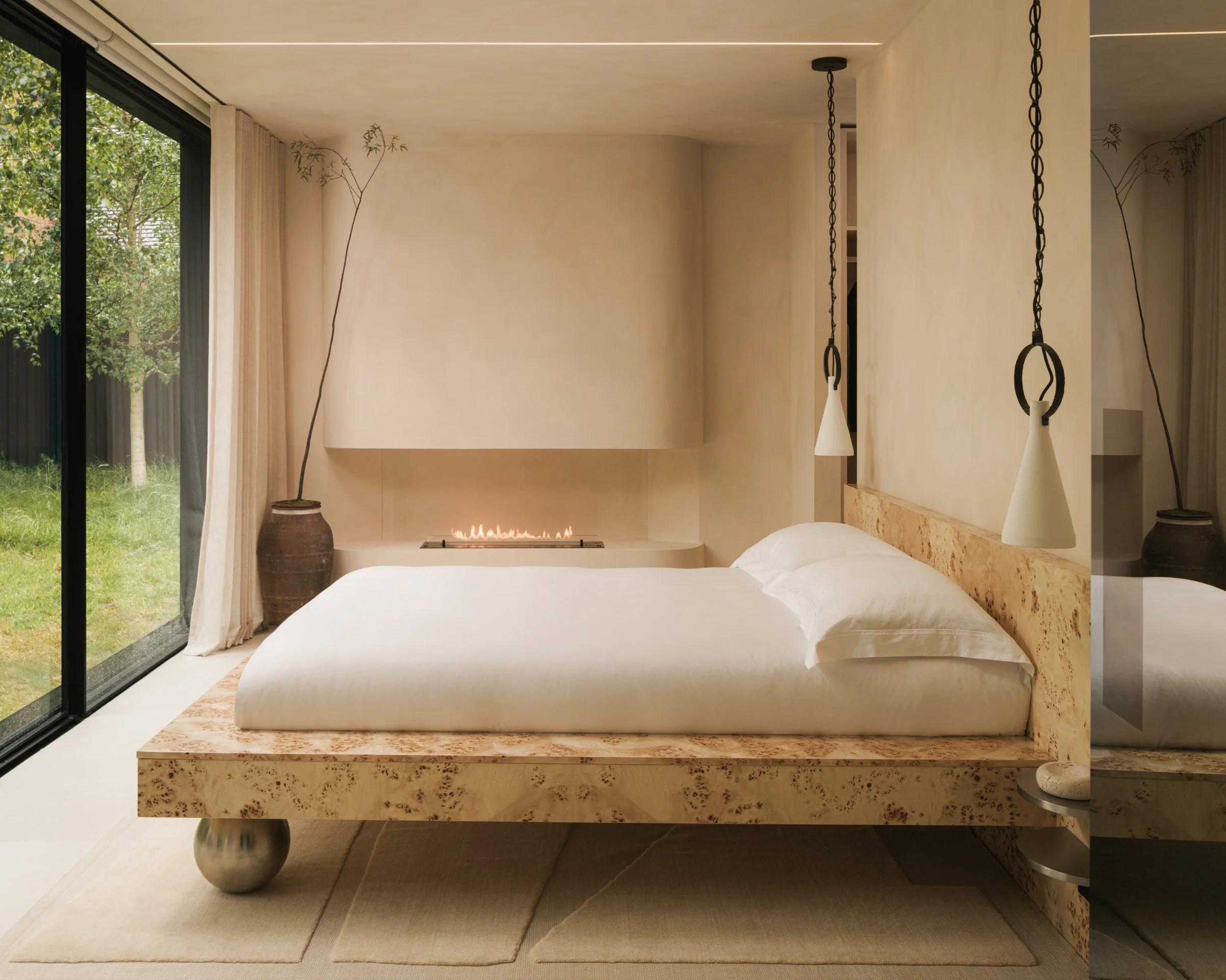 Minimalist bedroom with a bed featuring a natural wood headboard, white bedding, two pendant lamps hanging from the ceiling, a large floor-to-ceiling glass window with curtains, all painted in limewash