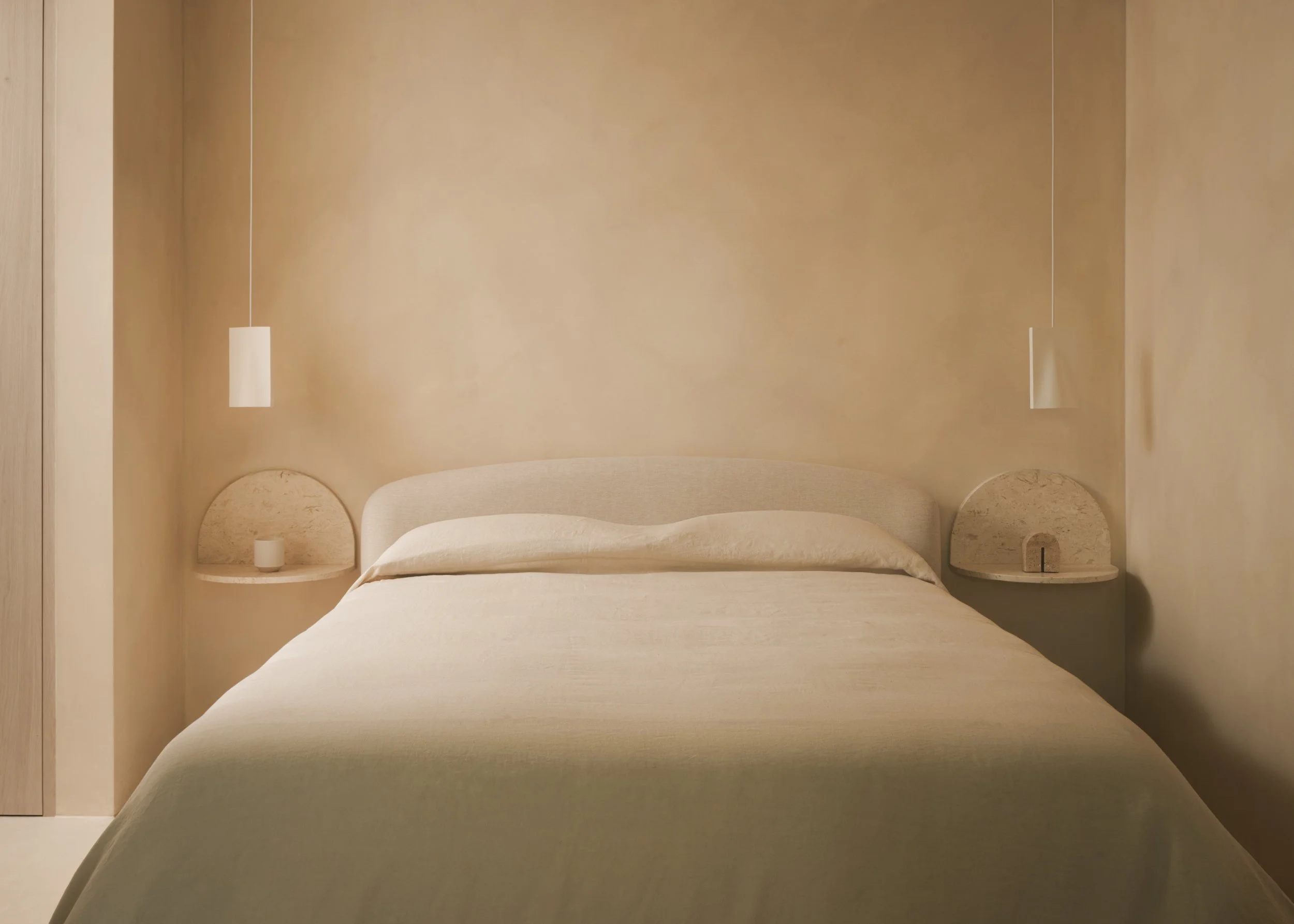 A minimalist bedroom finished in limewash and linen with a beige bed and matching headboard, two built-in round nightstands on either side, two hanging pendant lights, and a plain beige wall.