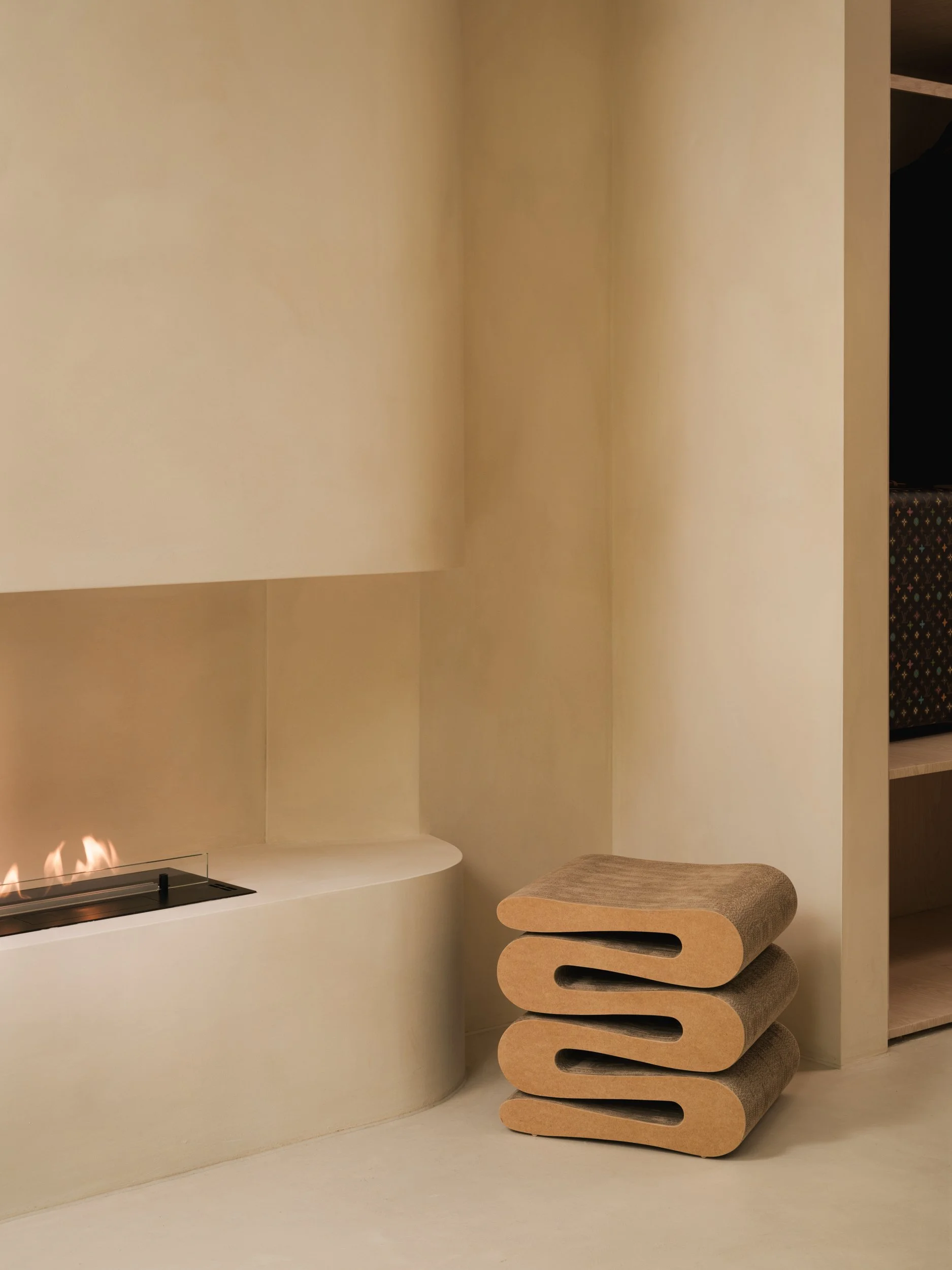 Minimalist interior painted with limewash with curved white built-in fireplace, beige stacked lounge chairs, and a bookshelf with patterned boxes.