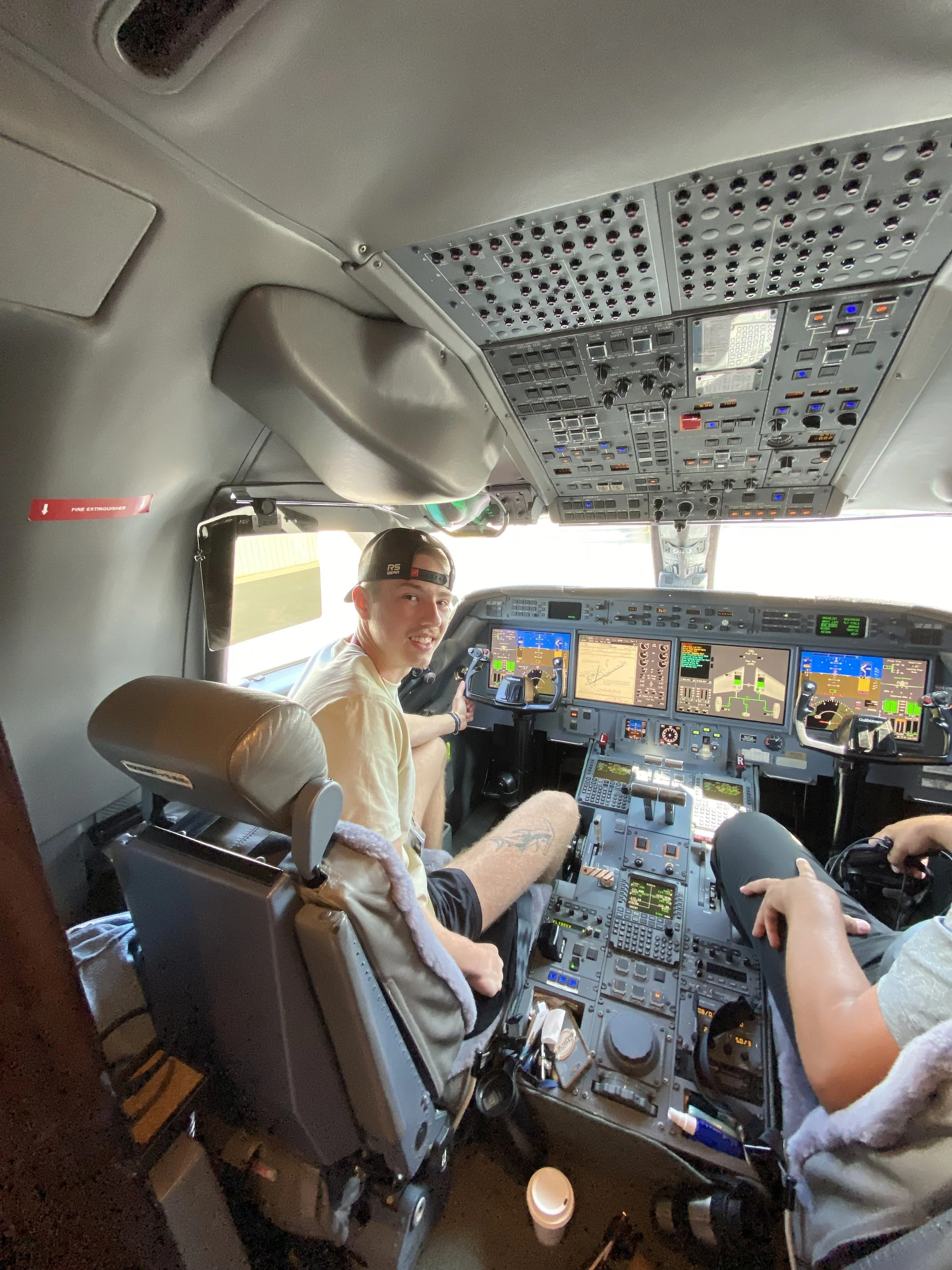 Interior of an airplane cockpit with two young men, one smiling at the camera, surrounded by various flight instruments and controls.