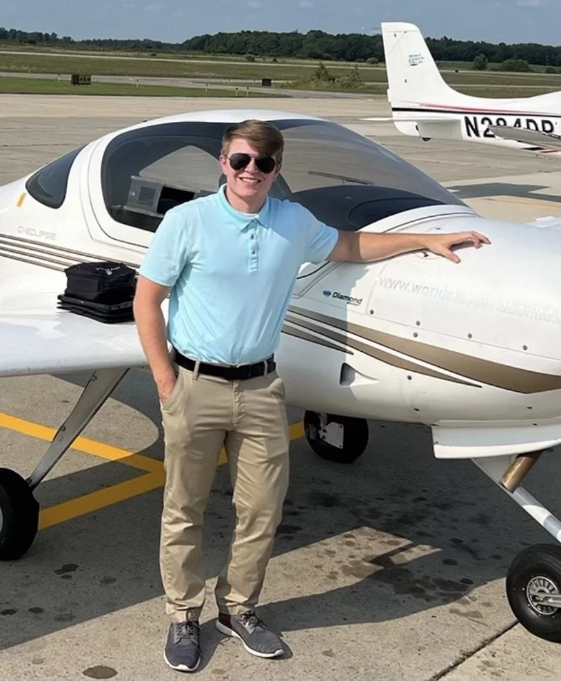 A smiling young man wearing sunglasses, a light blue polo shirt, and khaki pants standing next to a small white private airplane on an airport tarmac during the day.