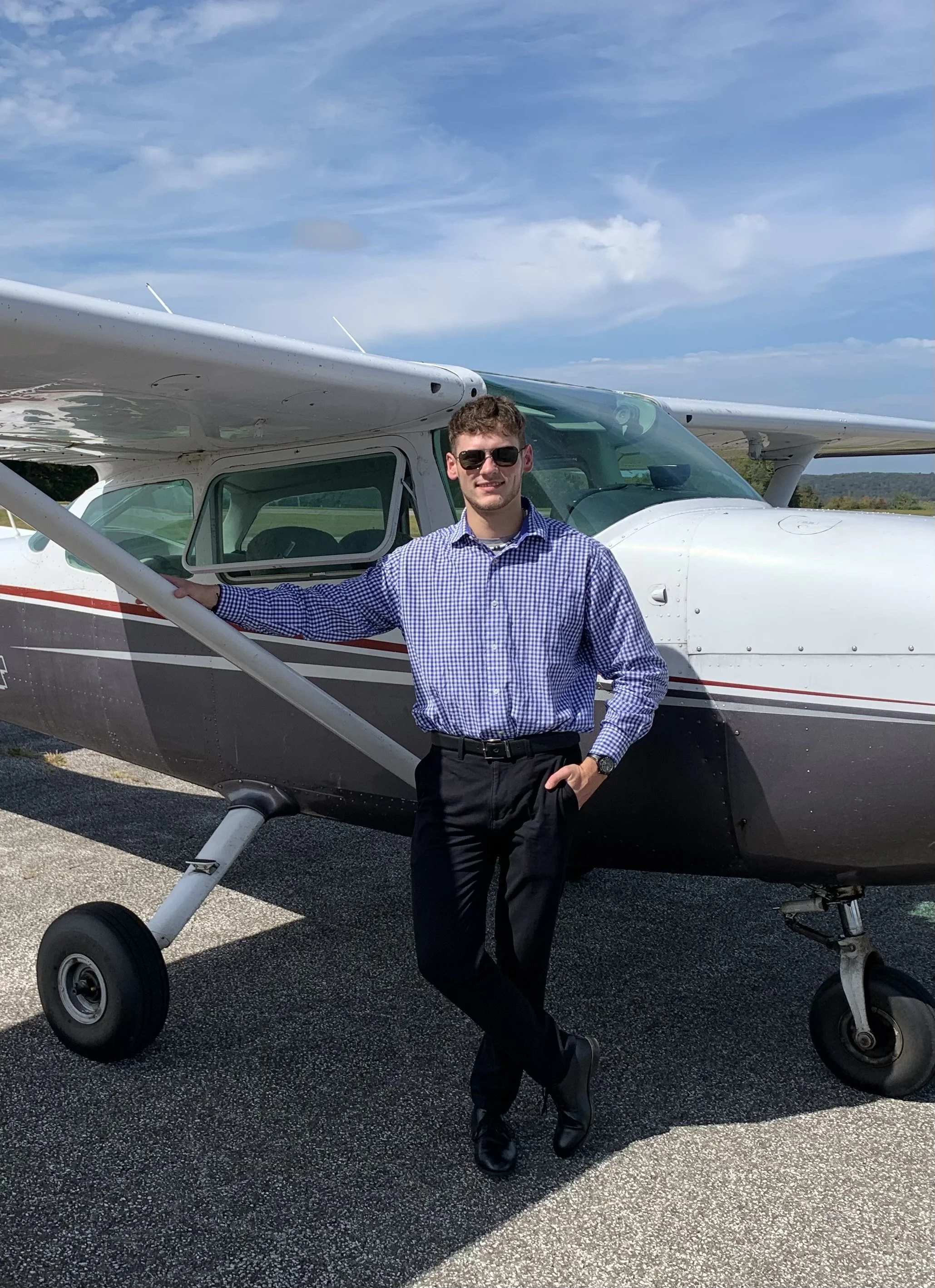 A man in sunglasses, a blue checkered shirt, and black pants standing next to a small white and black airplane on a sunny day.