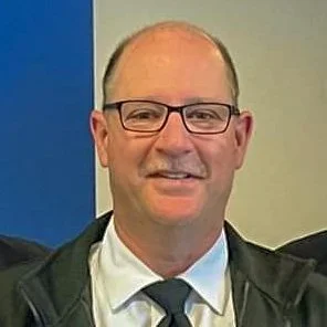 A middle-aged man with glasses, short hair, and a friendly smile, wearing a suit and tie, standing indoors against a neutral background.