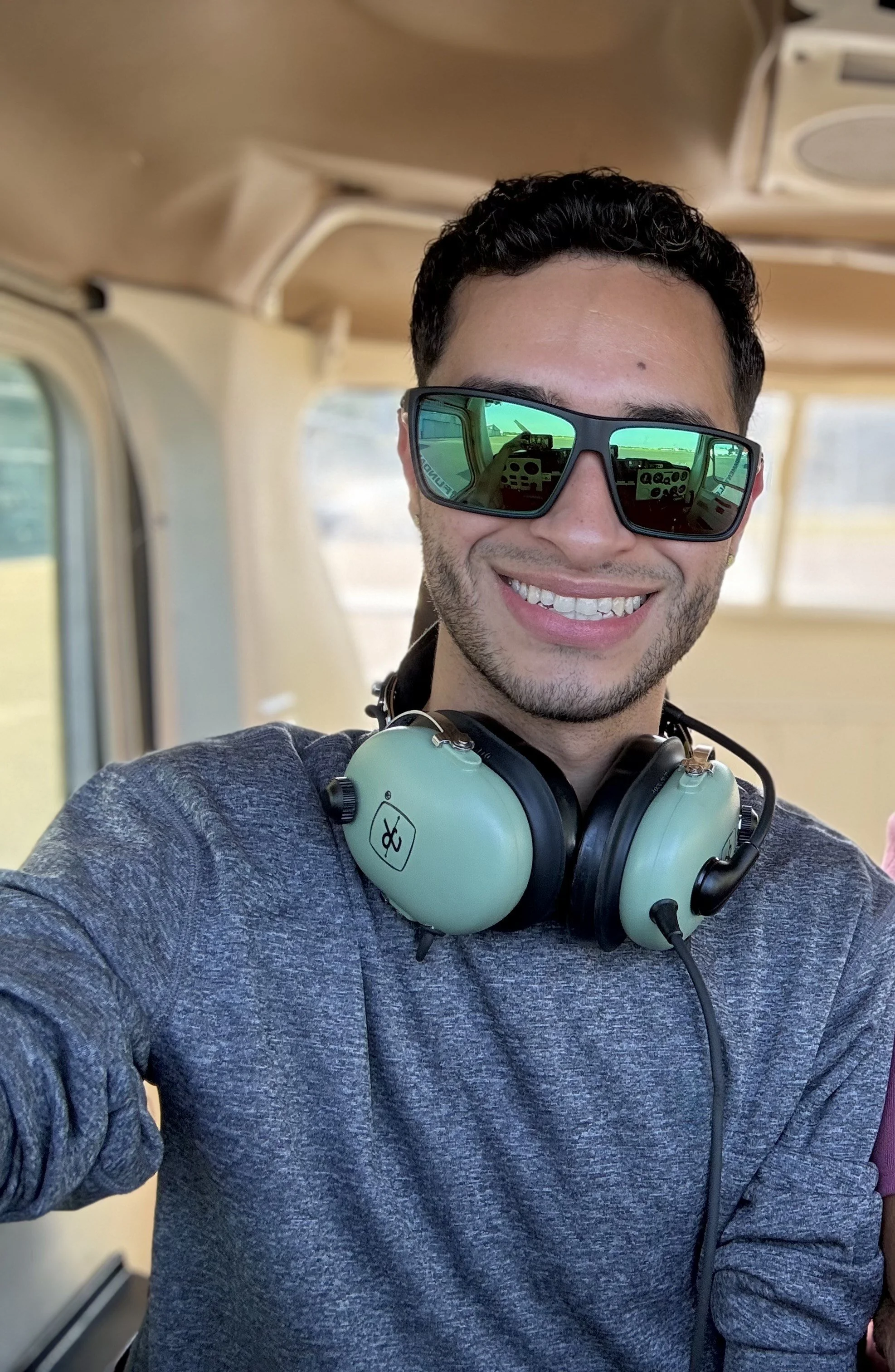 A smiling man wearing sunglasses and headphones takes a selfie inside a small aircraft or helicopter.