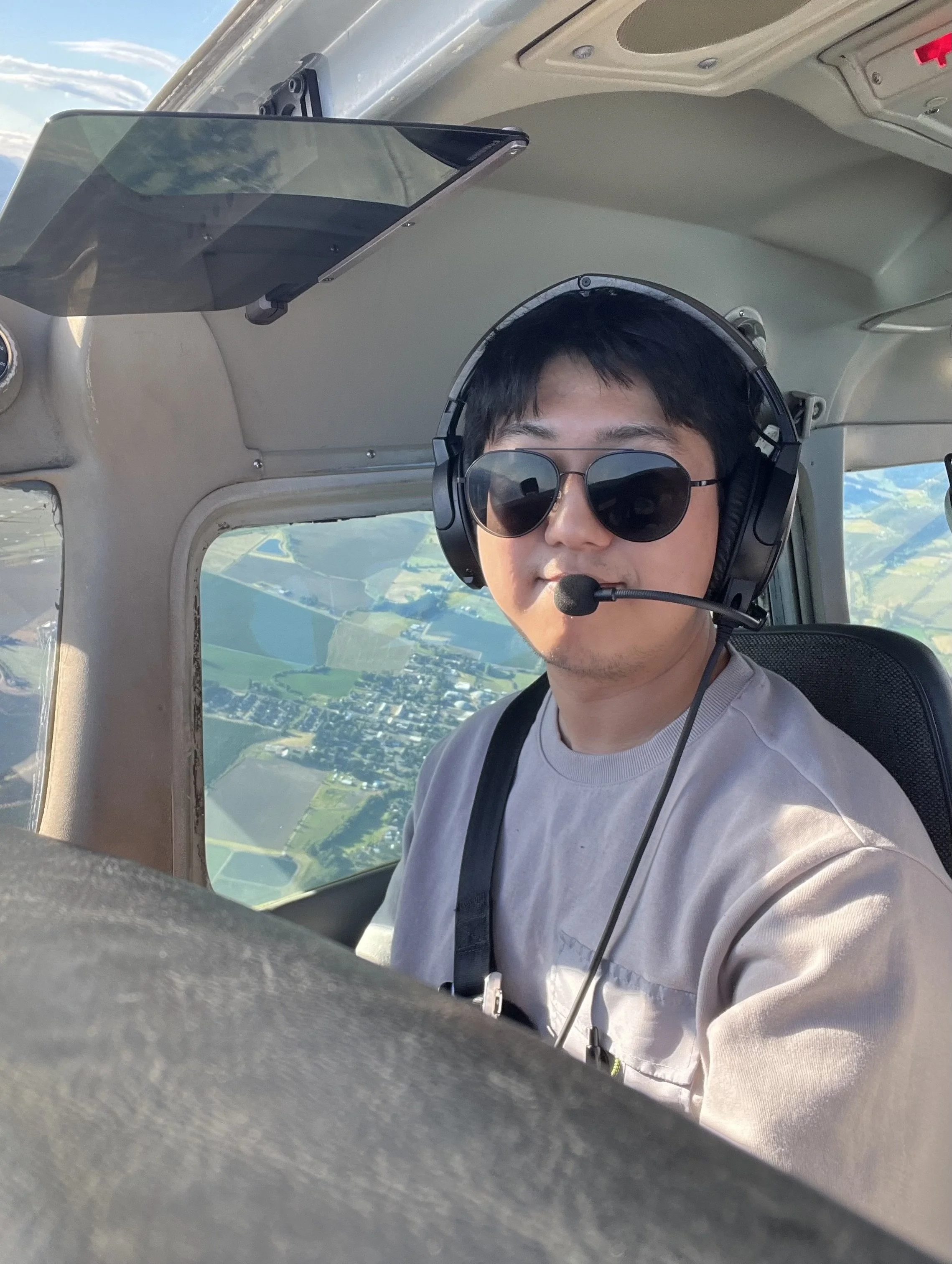 A man wearing sunglasses and a headset with a microphone is sitting in the cockpit of a small airplane, flying above a landscape of fields and a town.