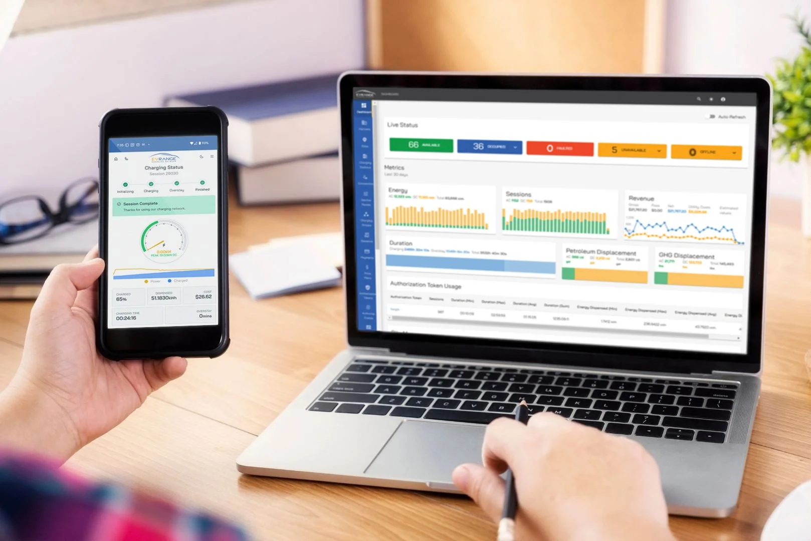Person holding a smartphone displaying a charging status app, working on a laptop showing analytics dashboards in a bright office setting with a desk, notebook, and plant nearby.