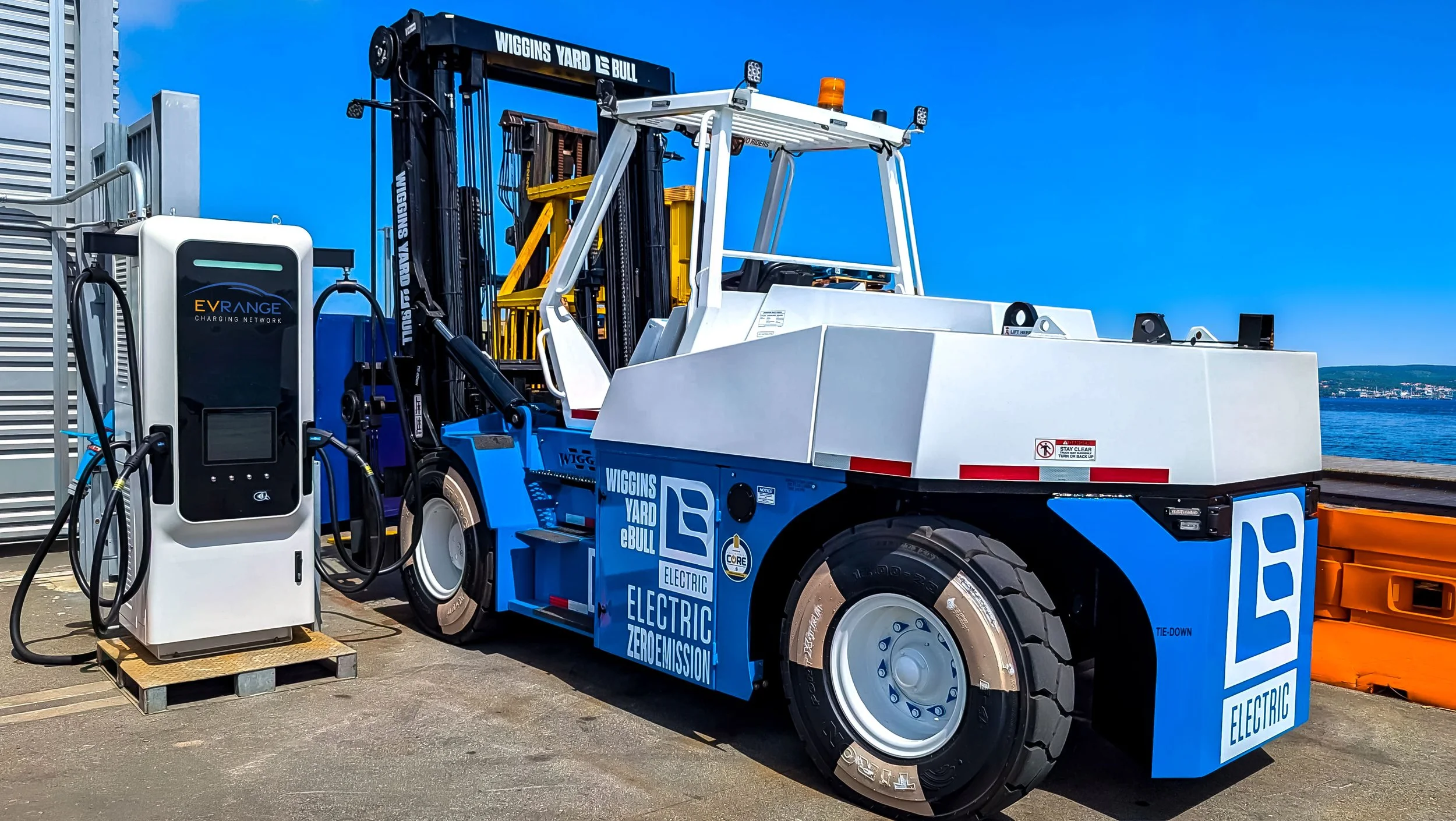 A white and blue Wiggins Yard eBull electric heavy-duty forklift is plugged into a dedicated white EV Range Charging Network DC fast charger at a sunny US port terminal, next to the waterfront.