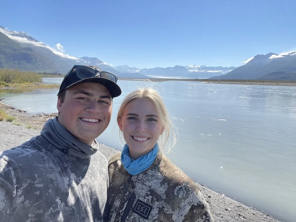 Two smiling people standing by a river with mountains and a clear blue sky in the background.