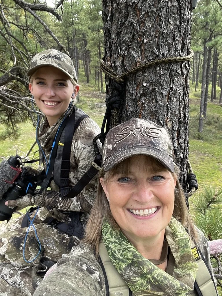Two women in camouflage attire smiling while sitting in a forested area, one holding binoculars and the other wearing a hat with the word 'STIHL' on it, near a tree.