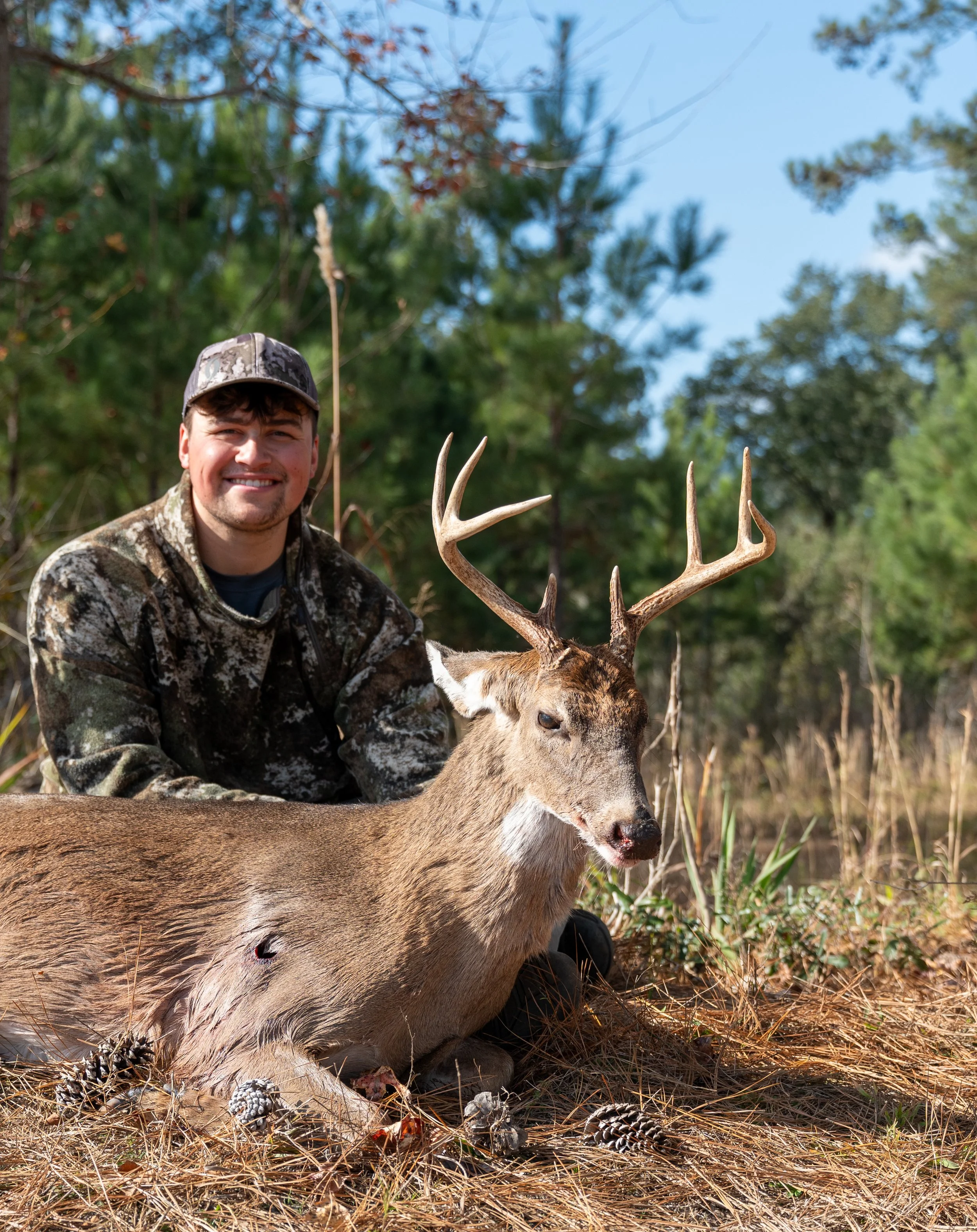 Two smiling people standing by a river with mountains and a clear blue sky in the background.