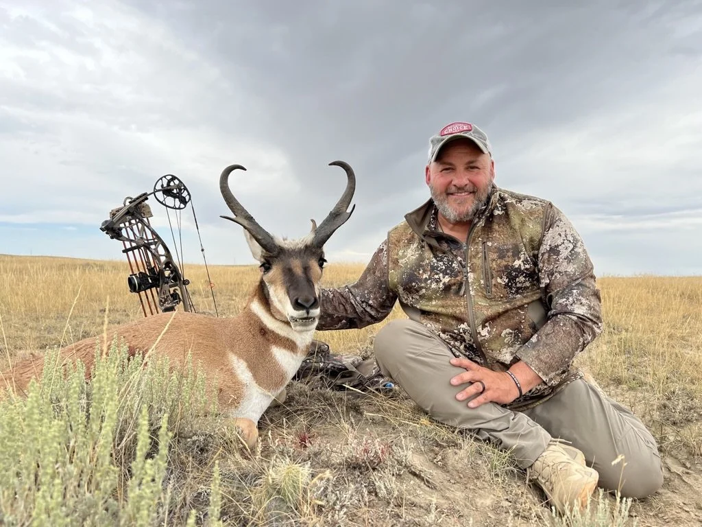 A man in camouflage clothing and a baseball cap sits next to a pronghorn antelope he has hunted, with a bow and arrow nearby, in an open grassy field with a cloudy sky.