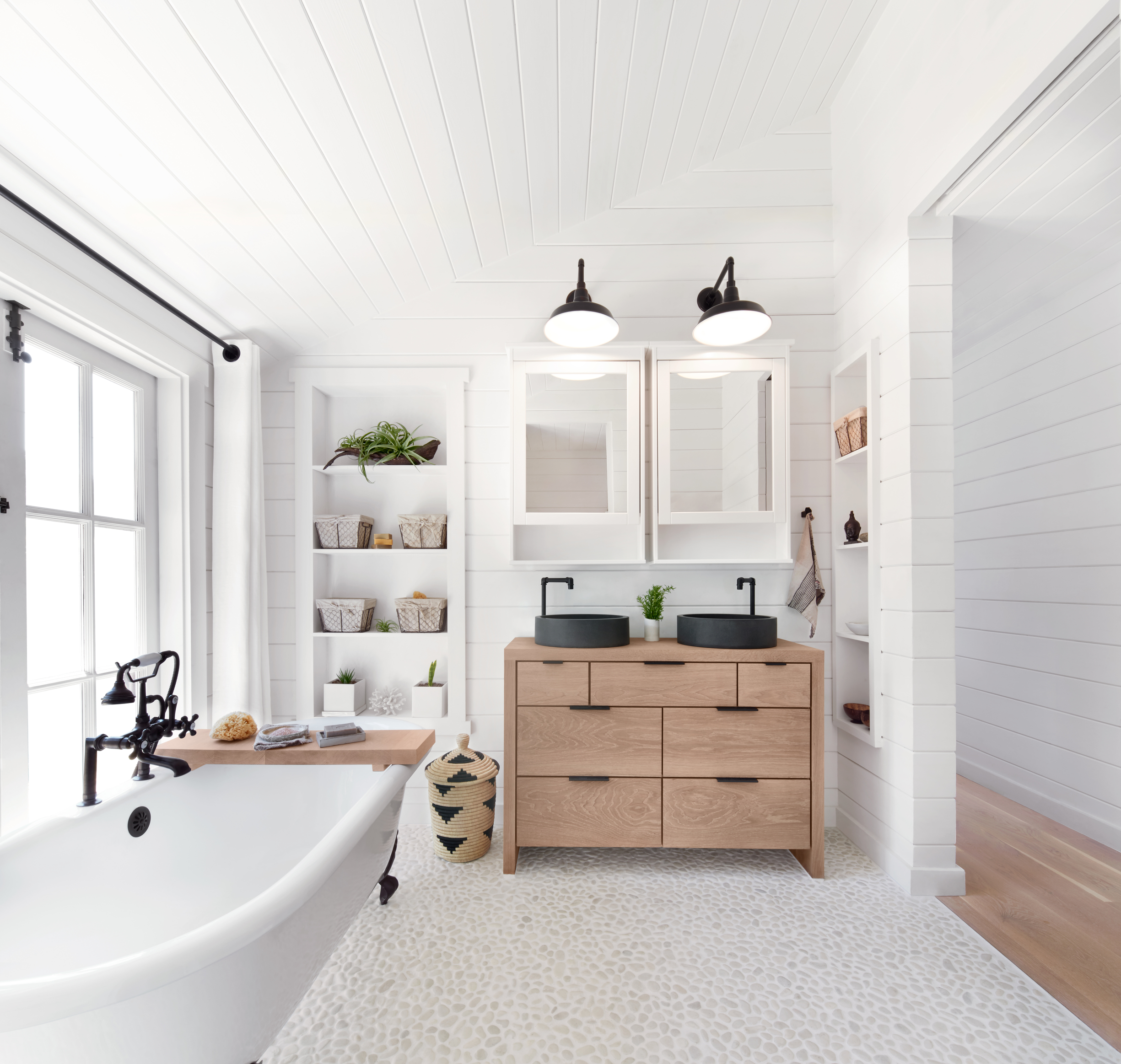 Modern bathroom interior with white freestanding bathtub, wooden vanity with double sinks, wall shelves with plants and baskets, and overhead lighting. Hermosa Beach, Venice, Mar Vista, Los Angeles, Manhattan Beach, Redondo Beach, South Bay Real Esta