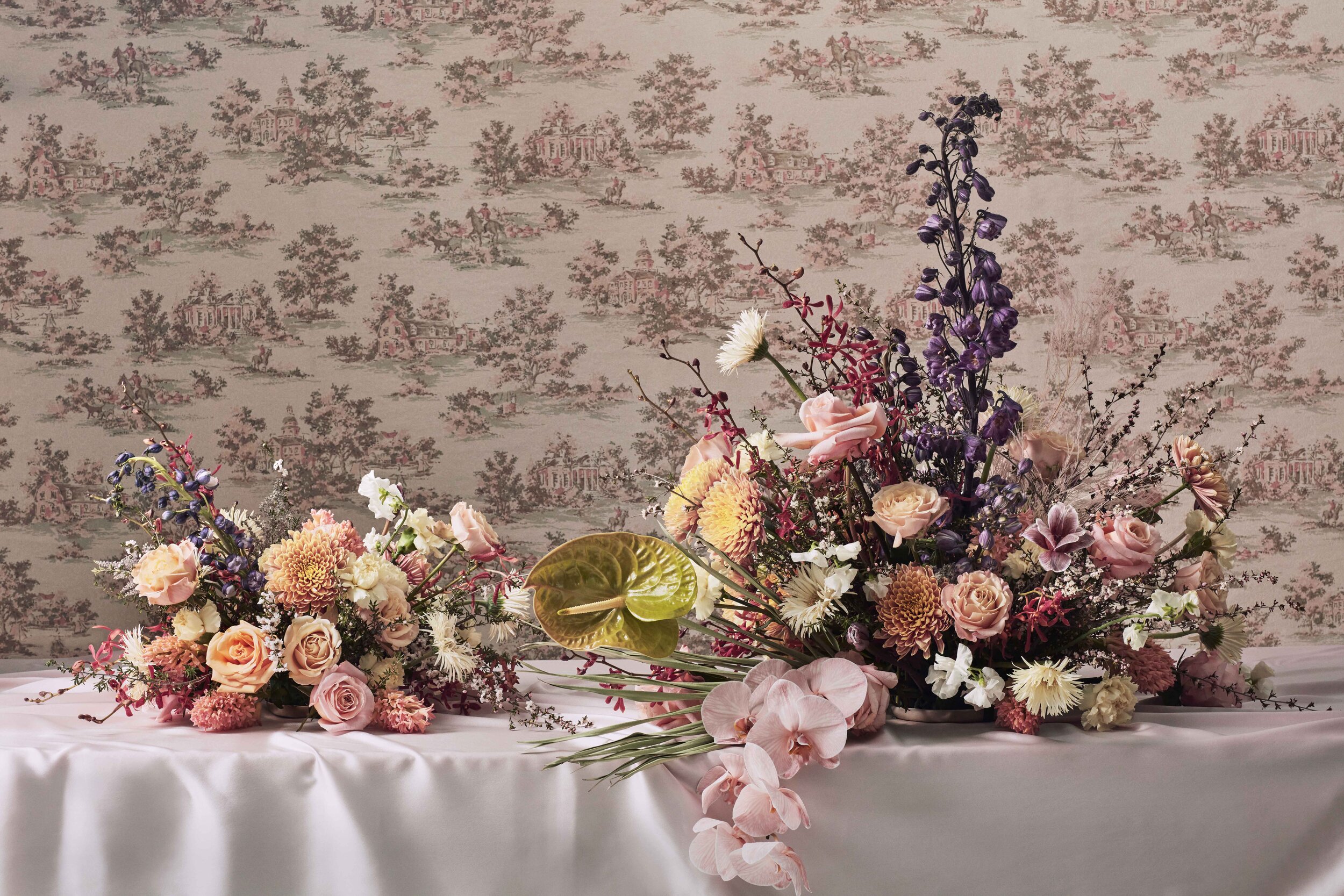 Two large floral arrangements with pink, purple, white, and peach flowers on a white draped table, with a wallpaper featuring a pastoral scene in the background.