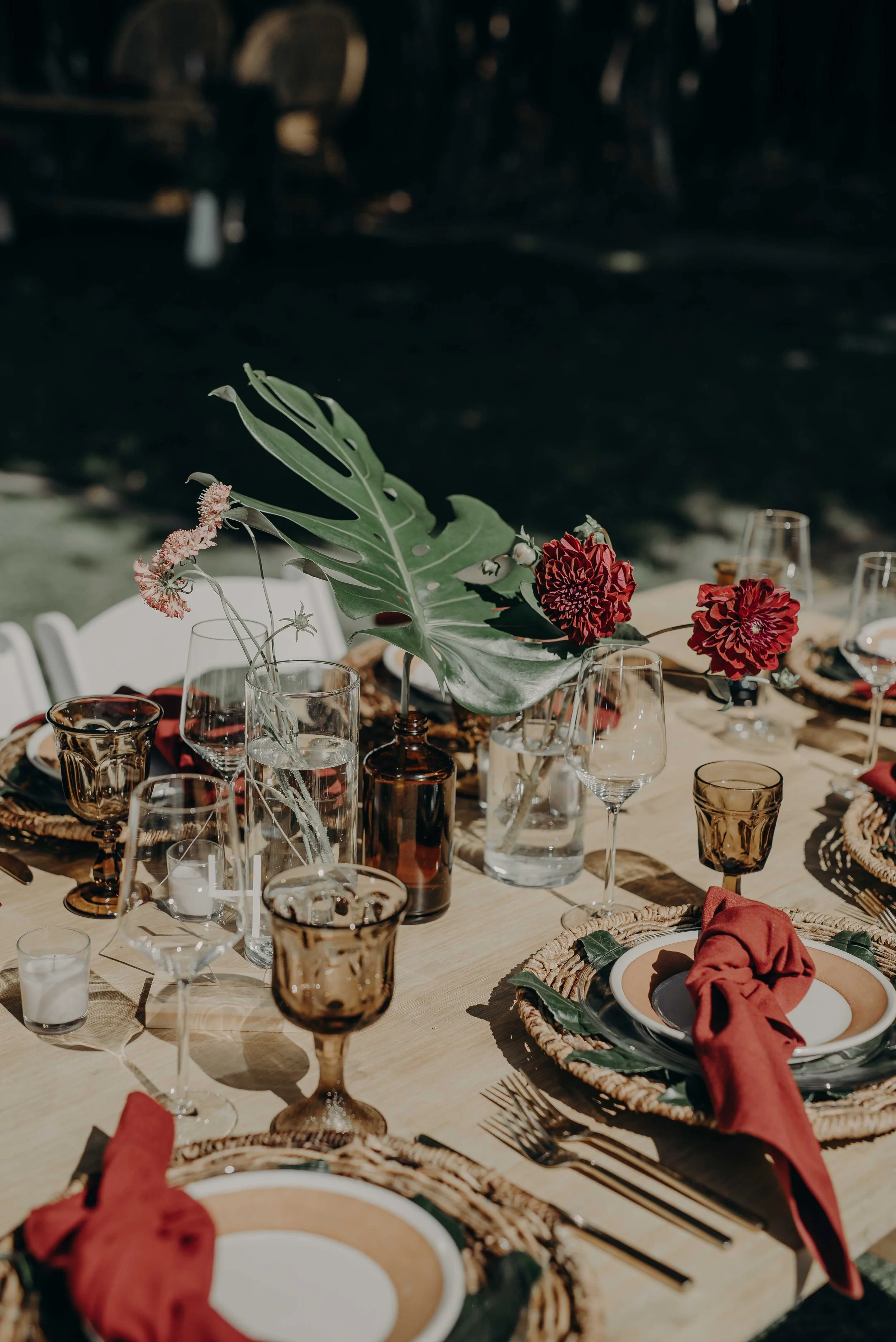 A rustic outdoor dining table decorated with a large floral centerpiece, wine glasses, candles, and set with plates, silverware, and red cloth napkins, illuminated by natural light.
