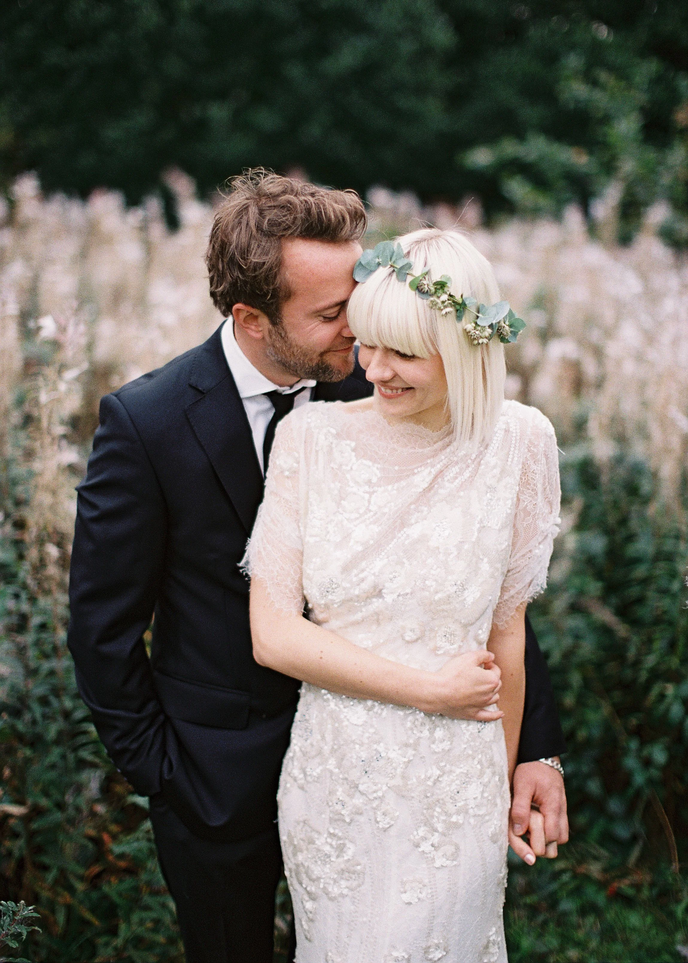 A couple in wedding attire smiling and embracing outdoors, with the man in a black tuxedo and the woman in a lace wedding dress and floral headpiece.