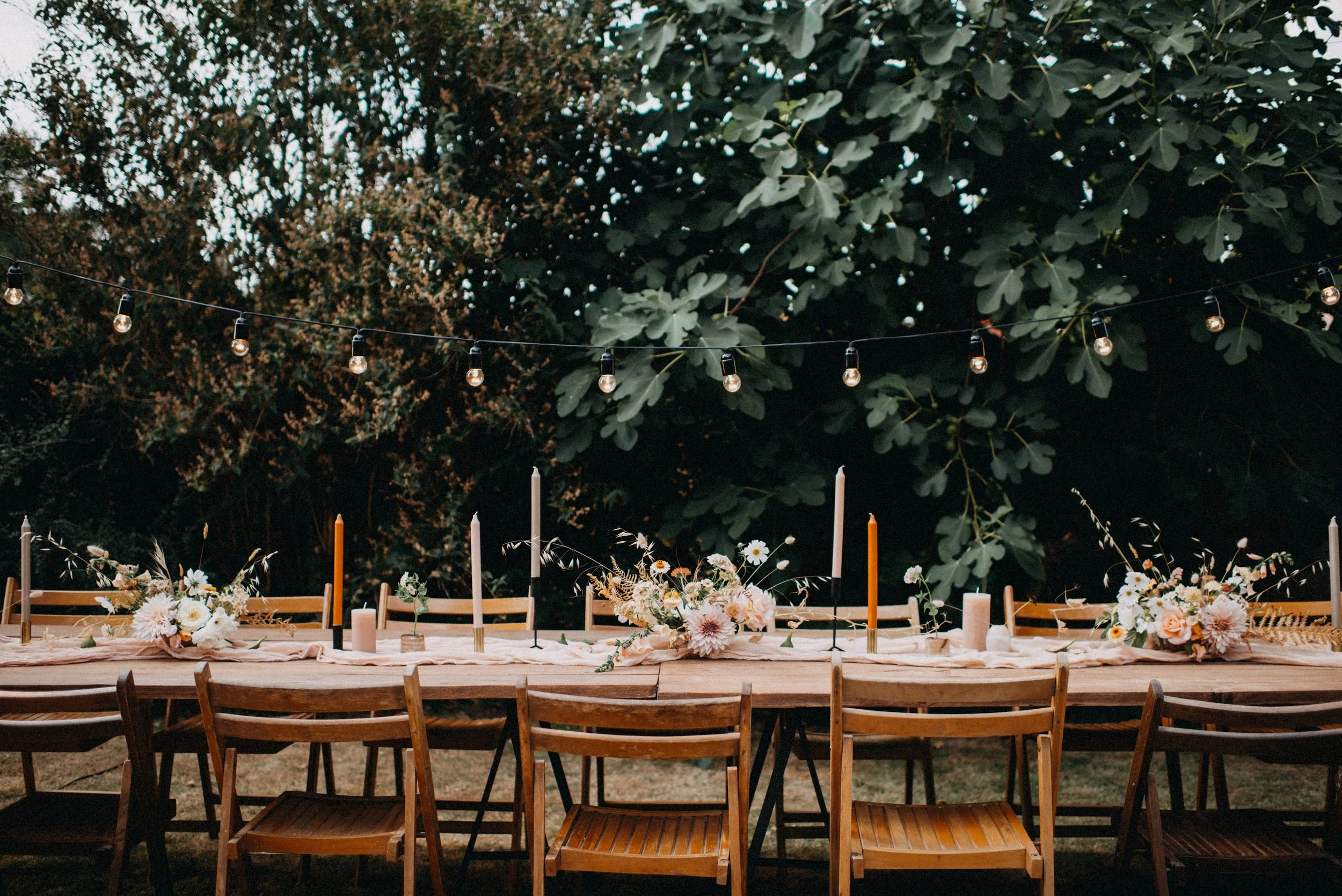 A long outdoor wooden table decorated with pink and white flowers, candles, and candles in various colors, set against a backdrop of dense greenery. String lights hang above the table.